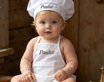 May include: A white chef hat and a matching white apron, both embroidered with the name Paular in black script. The apron features a front pocket and is worn by a baby sitting on a wooden table covered in flour, cookie shapes, and butter.