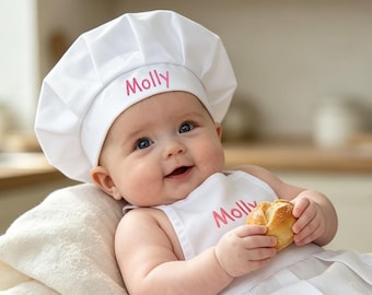 May include: A white chef hat and matching apron set featuring the name Molly embroidered in pink thread. The hat is pleated and the apron has a delicate white lace trim along the hem. The set is designed for a baby and includes the name Molly.