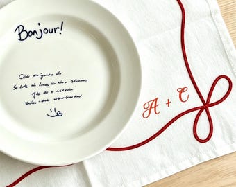 Puede incluir: Un mantel individual rectangular blanco con un borde bordado en rojo estilo cuerda con lazos en las esquinas y el monograma rojo A + C. Un plato de cerámica blanco con el texto Bonjour y escritura cursiva sobre el mantel con un tenedor.