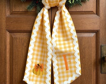 May include: A yellow and white gingham fabric ribbon with white scalloped edges, featuring an orange pumpkin graphic on one tail and the orange letter F on the other, attached to a green pine wreath with small red and green berries.