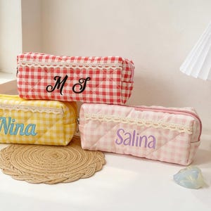 May include: Three rectangular pencil cases in red, yellow, and pink gingham patterns. Each case has a lace trim and embroidered text: "M J", "Nina", and "Salina". The cases are on a white surface near a window.
