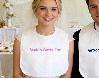 Puede incluir: Una novia y un novio con baberos blancos con texto. El babero de la novia dice "Bride's Gotta Eat" en rosa, y el del novio dice "Groom's Gotta Eat" en azul. Ambos están sentados a una mesa con una pequeña tarta sobre una servilleta.