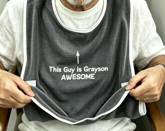 May include: A dark gray adult bib with white trim featuring an upward-pointing arrow and the white text "This Guy is Grayson AWESOME" printed on the front. The bib is made of a textured fabric and is worn over a white short-sleeved shirt.