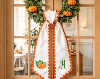 May include: A circular green foliage wreath adorned with six small orange pumpkins. A white fabric ribbon with a scalloped orange edge hangs from the center, featuring a green pumpkin illustration on one side and a green letter H on the other.