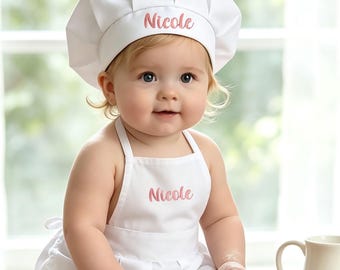 May include: A white chef hat and matching apron set featuring the name Nicole embroidered in pink script. The apron includes a delicate white lace trim along the bottom edge. The set is designed for a small child and is shown in a kitchen setting.