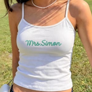 Puede incluir: Camiseta sin mangas blanca con el texto "Mrs. Simon" en verde. La camiseta se combina con vaqueros de mezclilla azul oscuro. Se ve un pequeño bolso blanco con una cadena plateada.