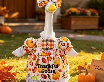May include: A white goose figurine wearing a dress and a matching bow, both featuring a pattern of pumpkins and autumn leaves. The dress has a plaid bib with two buttons, white lace trim, and the text Thankful Goose printed on the front.