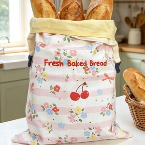 May include: A fabric bread bag with a light pink and white striped pattern featuring small colorful flowers and a red cherry graphic. The text "Fresh Baked Bread" is printed in red on the front. Three baguettes are inside the bag.