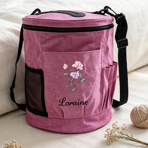 May include: A cylindrical mauve fabric bag with a black adjustable shoulder strap and black zipper closure. The front features a floral illustration in pink and purple with the name Loraine printed in black script, plus side mesh pockets.