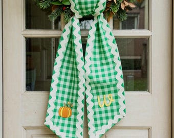May include: A green and white gingham fabric ribbon with a scalloped white edge, featuring an embroidered orange pumpkin on one side and a yellow cursive letter W on the other, hanging from a wreath made of pine needles and red berries.