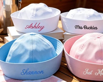 May include: Four sailor hats in white, light blue, and light pink. The white hats feature embroidered text reading Ashley and Mrs Buckles. The light blue hat features the name Shannon, and the light pink hat features the text Just Married.