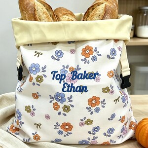 Pode incluir: Um saco de pão em tecido cor de creme com um padrão floral em tons de azul, laranja e verde. O texto "Top Baker Ethan" está bordado a azul na parte da frente. O saco tem uma bainha superior amarelo-claro e fivelas laterais de plástico preto.