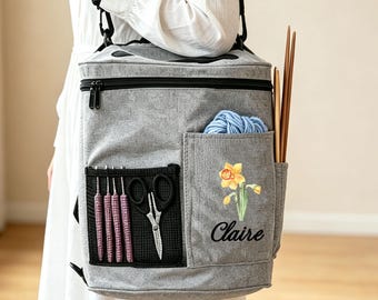 May include: A gray fabric craft bag with a black shoulder strap, featuring a mesh pocket containing four pink crochet hooks and scissors, a side pocket with blue yarn, a yellow daffodil graphic, and the name Claire printed in black script.