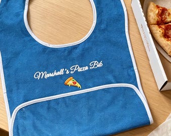 May include: A blue fabric bib with white trim featuring the embroidered text Marshall's Pizza Bib and a small pizza slice graphic. The bib has a curved neckline and a bottom pocket fold, designed for protection during meals.