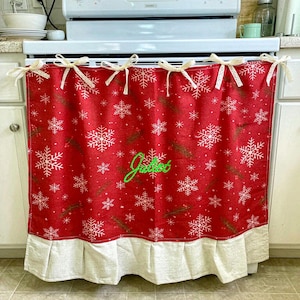 May include: A red fabric oven cover featuring a white snowflake and green pine needle pattern, personalised with the name Juliet in green script embroidery. The cover includes a cream-coloured ruffled bottom hem and five cream lace tie bows.