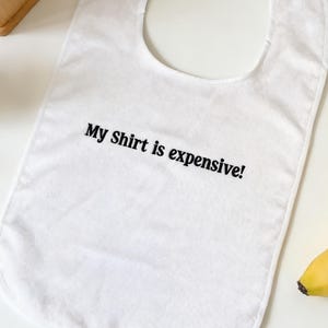 May include: White baby bib with the text "My Shirt is expensive!" in black. A yellow banana is next to the bib. A wooden box is in the background.
