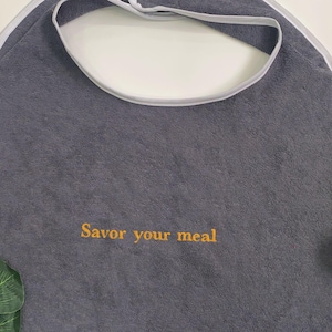 May include: A grey bib with white trim and the embroidered text "Savor your meal" in yellow. The bib is made of a soft material and is designed to catch food spills. A red apple and a green leaf are visible.