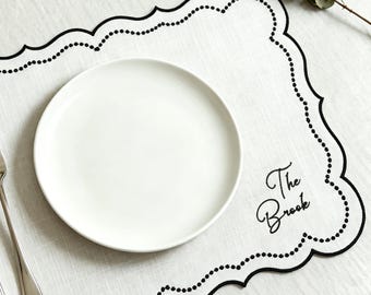 May include: A rectangular white linen placemat with a scalloped edge outlined in black embroidery and a dotted pattern. The text The Brook is embroidered in black script. A white ceramic plate, a silver fork, and a silver knife are placed on top.