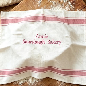 Op de afbeelding: Wit linnen keukendoek met twee horizontale rode strepen en de tekst "Annie Sourdough Bakery" in een cursief lettertype. De handdoek is opgevouwen op een houten snijplank met bloem.