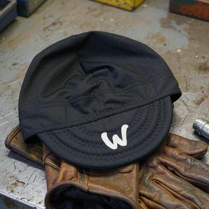 Puede incluir: Una gorra de ciclismo negra con una letra W blanca bordada en la visera, apoyada sobre un par de guantes de trabajo de cuero marrón desgastados. Un pequeño componente de herramienta cilíndrico de metal plateado se sitúa a la derecha.
