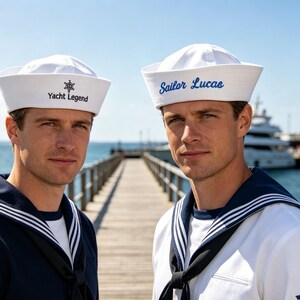 Op de afbeelding: Twee matrozenpetten, één wit met de zwarte tekst Yacht Legend en een icoon van een scheepsroer, en één wit met de blauwe cursieve tekst Sailor Lucas. Beide petten hebben een traditionele platte bovenkant en een brede, omgevouwen rand.