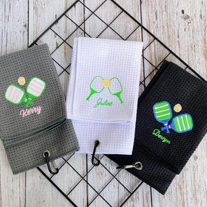 May include: Three waffle-weave golf towels in gray, white, and black, each embroidered with a pickleball design and a name. The gray towel has pink accents and the name "Kerry". The white towel has green accents and the name "Juliet". The black towel has blue accents and the name "Derryn".