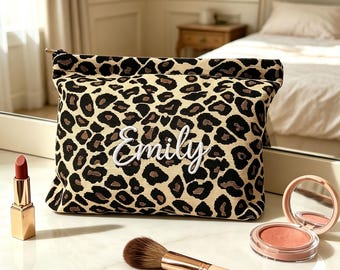 May include: A rectangular zippered pouch featuring a tan and black leopard print pattern with the name Emily embroidered in white script on the front. The pouch is accompanied by a gold-cased lipstick, a makeup brush, and a compact mirror.