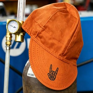May include: A solid orange welding cap featuring a black embroidered skeleton hand making a rock and roll sign on the front brim. The cap is constructed with visible stitched seams and a rounded crown, designed for head protection during welding tasks.
