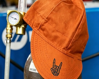 May include: A solid orange welding cap featuring a black embroidered skeleton hand making a rock and roll sign on the front brim. The cap is constructed with visible stitched seams and a rounded crown, designed for head protection during welding tasks.