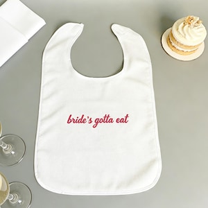 Puede incluir: Babero blanco con el texto "bride's gotta eat" en escritura roja. La imagen también muestra una servilleta blanca doblada, dos copas de champán y un pastel pequeño con glaseado blanco.