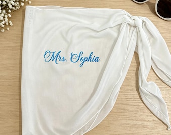 Puede incluir: Un pareo blanco con el texto bordado Mrs. Sophia en letras cursivas azules. La tela es ligera y semitransparente, diseñada con un cierre de nudo lateral. La prenda se muestra junto a unas gafas de sol blancas y flores de paniculata.
