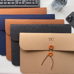 Op de afbeelding: Een verzameling vilten laptophulzen in verschillende kleuren: beige, marineblauw, bruin en antracietgrijs. De voorste hoes heeft de initialen "TC" en houten knoopsluitingen. Een toetsenbord, glas en telefoon zijn op de achtergrond te zien.