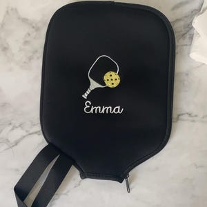 Puede incluir: Funda negra para raqueta de pickleball con cierre de cremallera y asa. La funda presenta un diseño bordado de raqueta y pelota de pickleball en blanco y amarillo, con el nombre "Emma" bordado debajo.