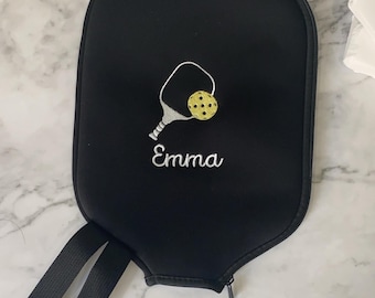 Puede incluir: Funda negra para raqueta de pickleball con cierre de cremallera y asa. La funda presenta un diseño bordado de raqueta y pelota de pickleball en blanco y amarillo, con el nombre "Emma" bordado debajo.