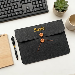 Puede incluir: Una funda para portátil de fieltro gris oscuro con el nombre "Susie" bordado en amarillo. La funda tiene dos botones de madera y un cierre de cordón marrón. También se ven un teclado negro, una planta suculenta, un bloc de notas, un bolígrafo y una taza.