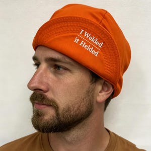 Puede incluir: Una gorra de soldador de color naranja brillante que presenta un ala ancha y doblada con costuras en zigzag visibles. El texto "I Welded it Helded" está impreso en blanco en el lateral del ala. La gorra está hecha de tela de color sólido.