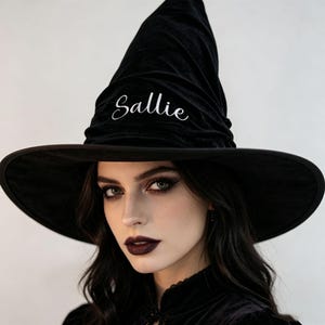 May include: A black velvet witch hat featuring a tall, pointed crown with a wide brim. The word Sallie is embroidered in white cursive script on the front of the crown. The hat has a soft, textured appearance with subtle folds in the fabric.