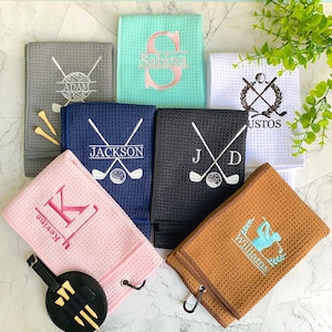 Puede incluir: Variedad de toallas de golf personalizadas en varios colores, incluyendo gris, azul, negro, rosa, blanco y marrón. Cada toalla presenta diseños bordados con nombres, iniciales o gráficos temáticos de golf. Algunas toallas tienen un lazo y un soporte para tees.