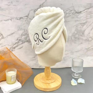 May include: A cream-coloured microfibre hair towel wrap featuring the embroidered black monogram letters RC on the side. The towel is draped over a beige mannequin head stand, showcasing a soft, plush texture and a gathered, turban-style design.