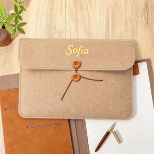Puede incluir: Una funda para portátil de fieltro beige con el nombre "Sofia" en letras doradas. La funda tiene un cierre de solapa asegurado por dos botones de madera y una cuerda. También se ven un cuaderno de cuero marrón y un bolígrafo.