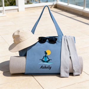 Könnte beinhalten: Hellblaue Tragetasche mit Yogamatte, Sonnenbrille, beigem Sonnenhut und grauer Leggings. Die Tasche hat eine Yoga-Grafik und den Namen "Ashely". Die Gegenstände sind auf einer gefliesten Oberfläche angeordnet.