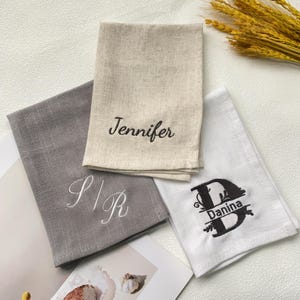 Pode incluir: Três toalhas de linho dobradas em cinza, bege e branco. A toalha bege tem "Jennifer" bordado em preto. A toalha cinza tem "S|R" bordado em branco. A toalha branca tem "D Danina" bordado em preto.