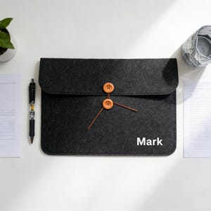 Puede incluir: Una funda para portátil de fieltro gris oscuro con cierre de solapa, asegurada con dos botones naranjas y una cuerda marrón. El nombre "Mark" está impreso en blanco. Se ven un bolígrafo y una planta.