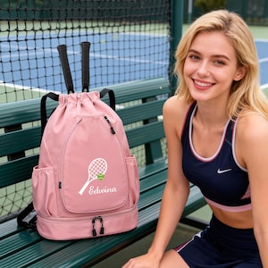 Può includere: Uno zaino da tennis rosa con chiusura a coulisse e spallacci neri. La borsa presenta una grafica di racchetta da tennis bianca con il nome "Edwina" e una pallina da tennis. Sono visibili due racchette da tennis. La borsa ha tasche laterali e un vano inferiore con cerniera.
