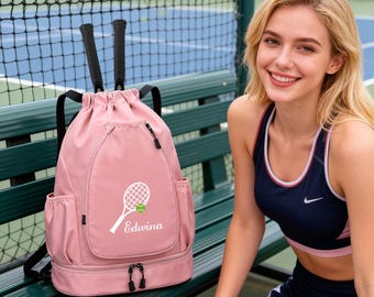 Puede incluir: Una mochila de tenis rosa con cierre de cordón y correas negras. La bolsa presenta un gráfico de raqueta de tenis blanca con el nombre "Edwina" y una pelota de tenis. Se ven dos raquetas de tenis. La bolsa tiene bolsillos laterales y un compartimento inferior con cremallera.