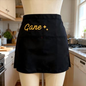 Puede incluir: Delantal de cintura negro con el nombre "Cane" bordado en oro, acompañado de dos estrellas doradas. El delantal tiene un bolsillo y una costura roja. El delantal se muestra en un maniquí en un entorno de cocina.
