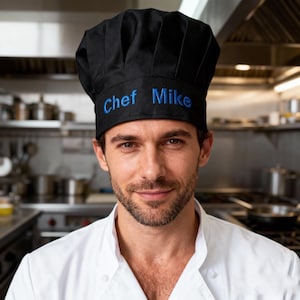 May include: A black chef's hat with the embroidered text "Chef Mike" in blue. The person is wearing a white chef's coat. The background is a commercial kitchen, with stainless steel appliances.