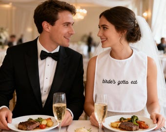 Puede incluir: Escena de boda con una pareja en una mesa. La novia lleva un babero blanco con el texto "birde's gotta eat". El novio lleva un esmoquin negro. En la mesa hay platos de comida y copas de champán. Una rosa está en primer plano.