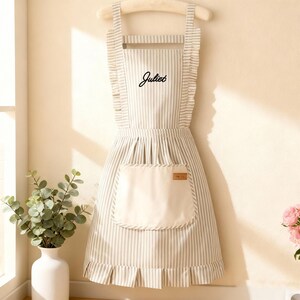 May include: A striped apron with the name "Juliet" in black script. The apron has a large front pocket and ruffled edges. The apron is hanging on a wooden hanger.