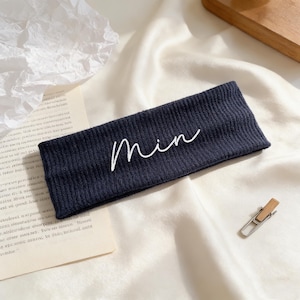May include: A navy blue headband with the cursive word "Min" embroidered in white. The headband is made of a textured knit fabric and is displayed on a white, textured surface.